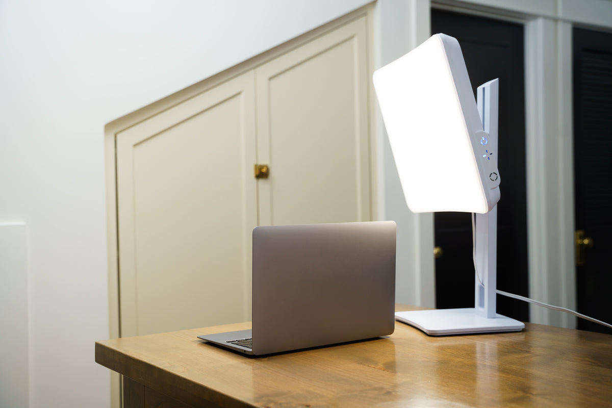 Laptop on a wooden desk with a large light therapy lamp in a room.