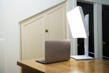 Laptop on a wooden desk with a large light therapy lamp in a room.