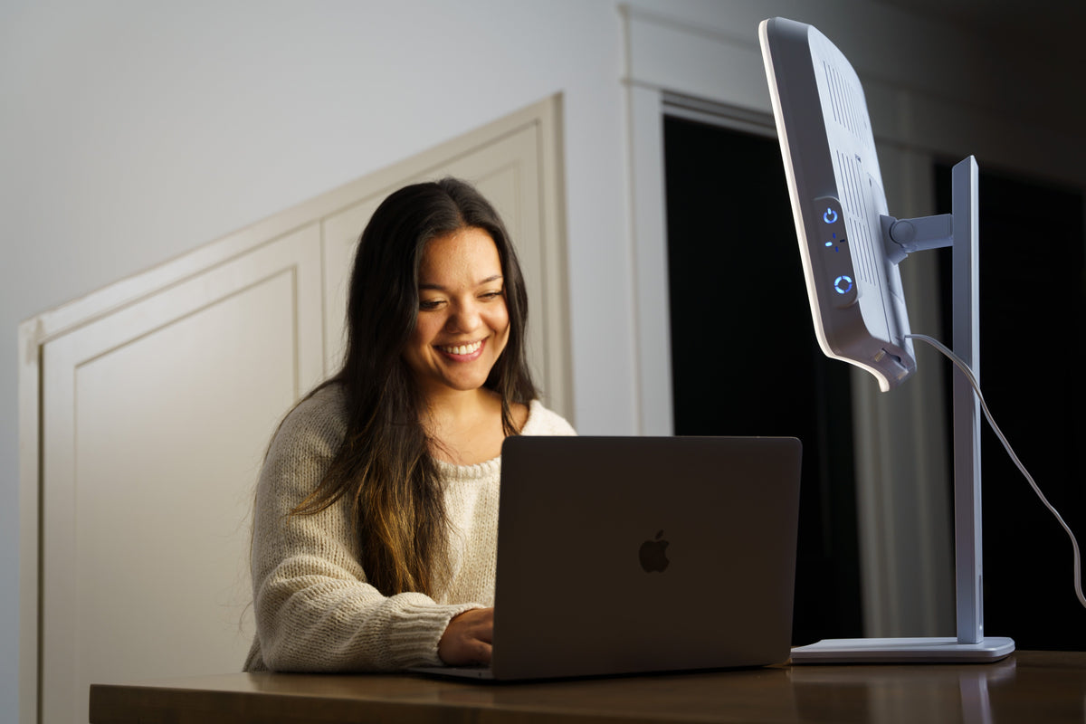 A woman smiling while working on a laptop at a desk, with a white light therapy lamp sitting next to her.