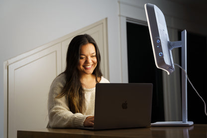 A woman smiling while working on a laptop at a desk, with a white light therapy lamp sitting next to her.